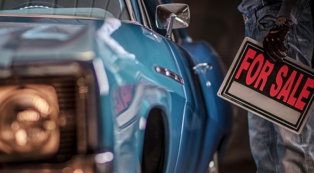 A person holding a "For Sale" sign to sell my car near a blue car.