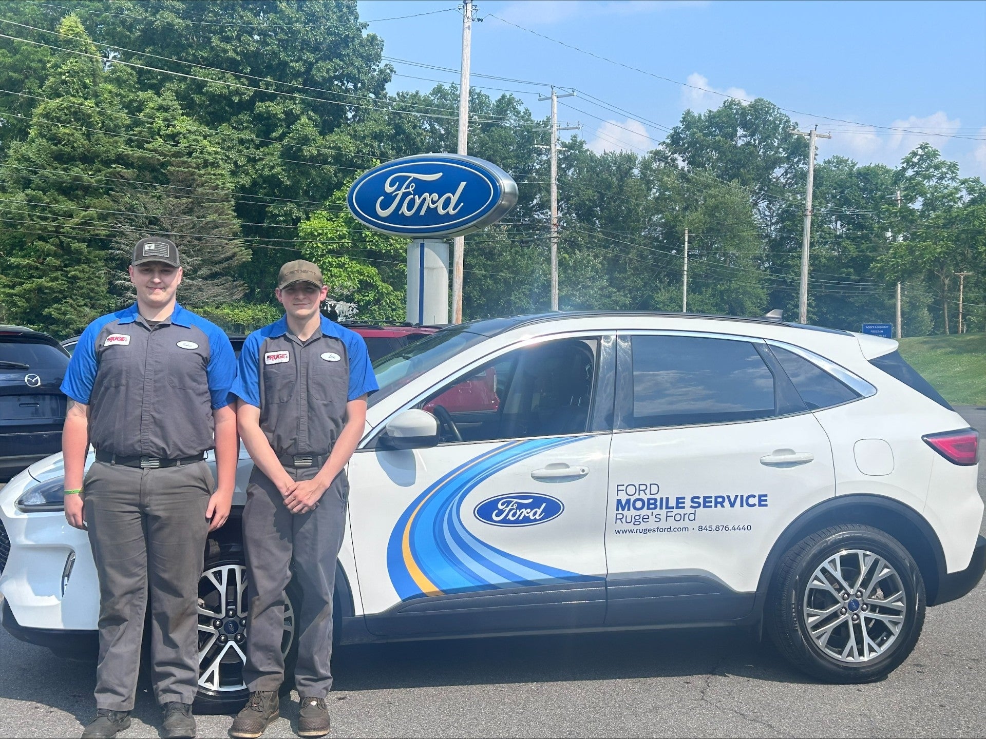 Two young men in uniforms stand next to a white Ford Mobile Service vehicle under a Ford sign.