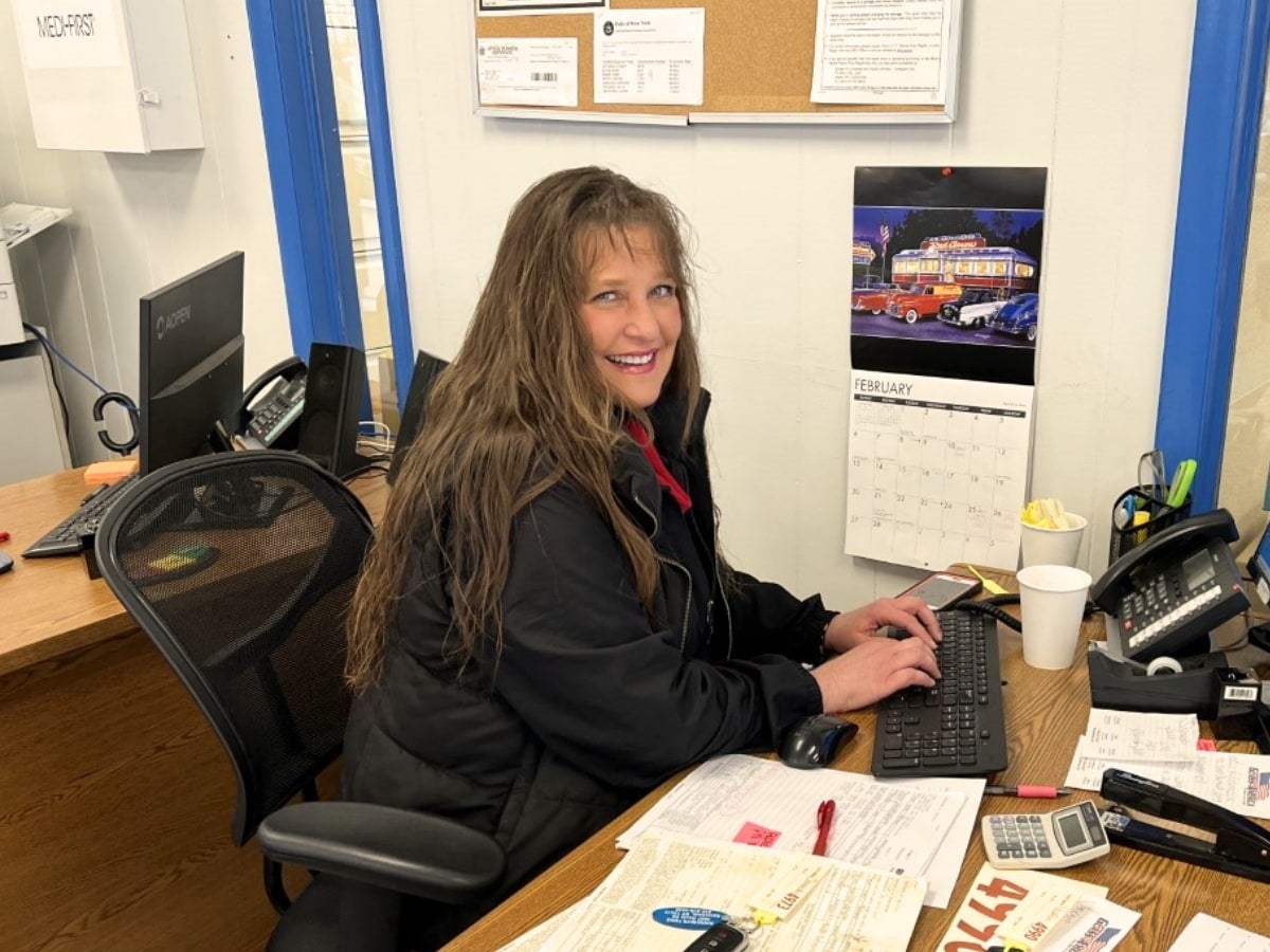 A woman with long brown hair smiles at her office desk with a calendar, computer, and phone.