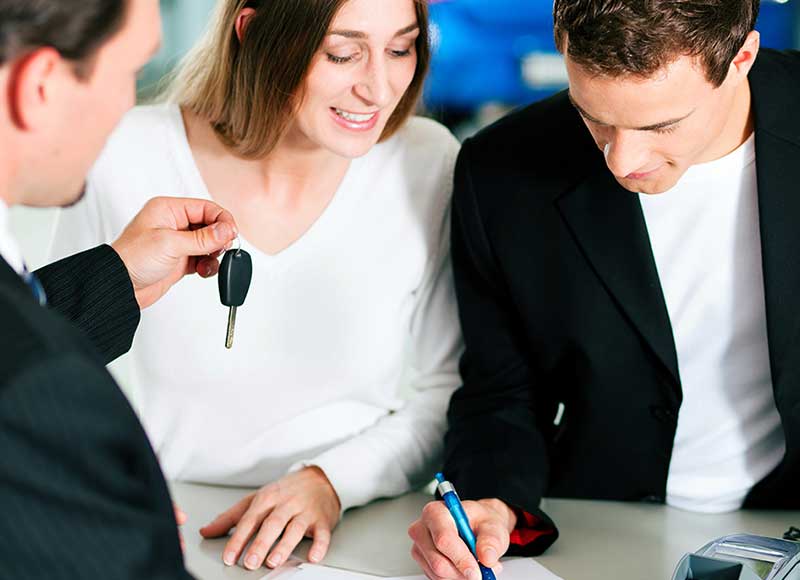 A salesperson hands car keys to a smiling woman as a man signs paperwork.