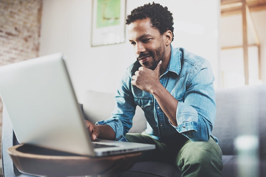 A smiling man with a beard sits on a sofa, looking at a laptop open on a small table.