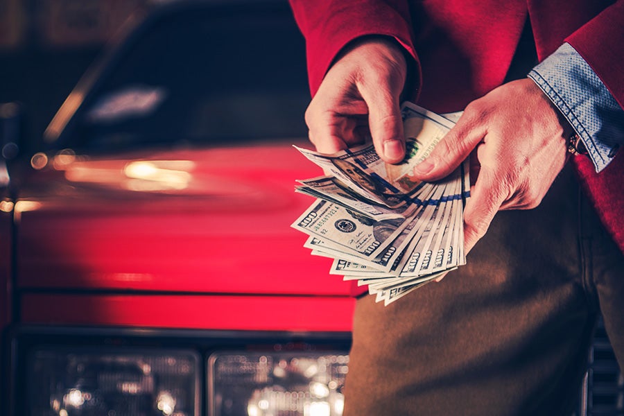 Hands holding a large fan of hundred-dollar bills, with a red car partially visible in the background.